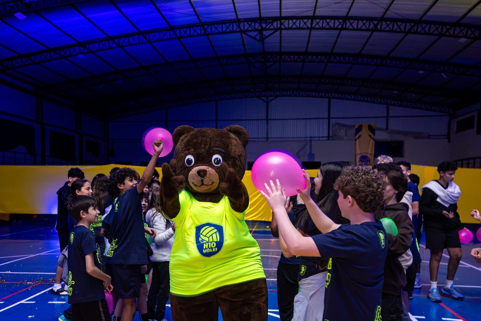 Quadra coberta a noite sob luz azul: acampantes em volta do mascote do camp, com baloes rosa e verde no alto