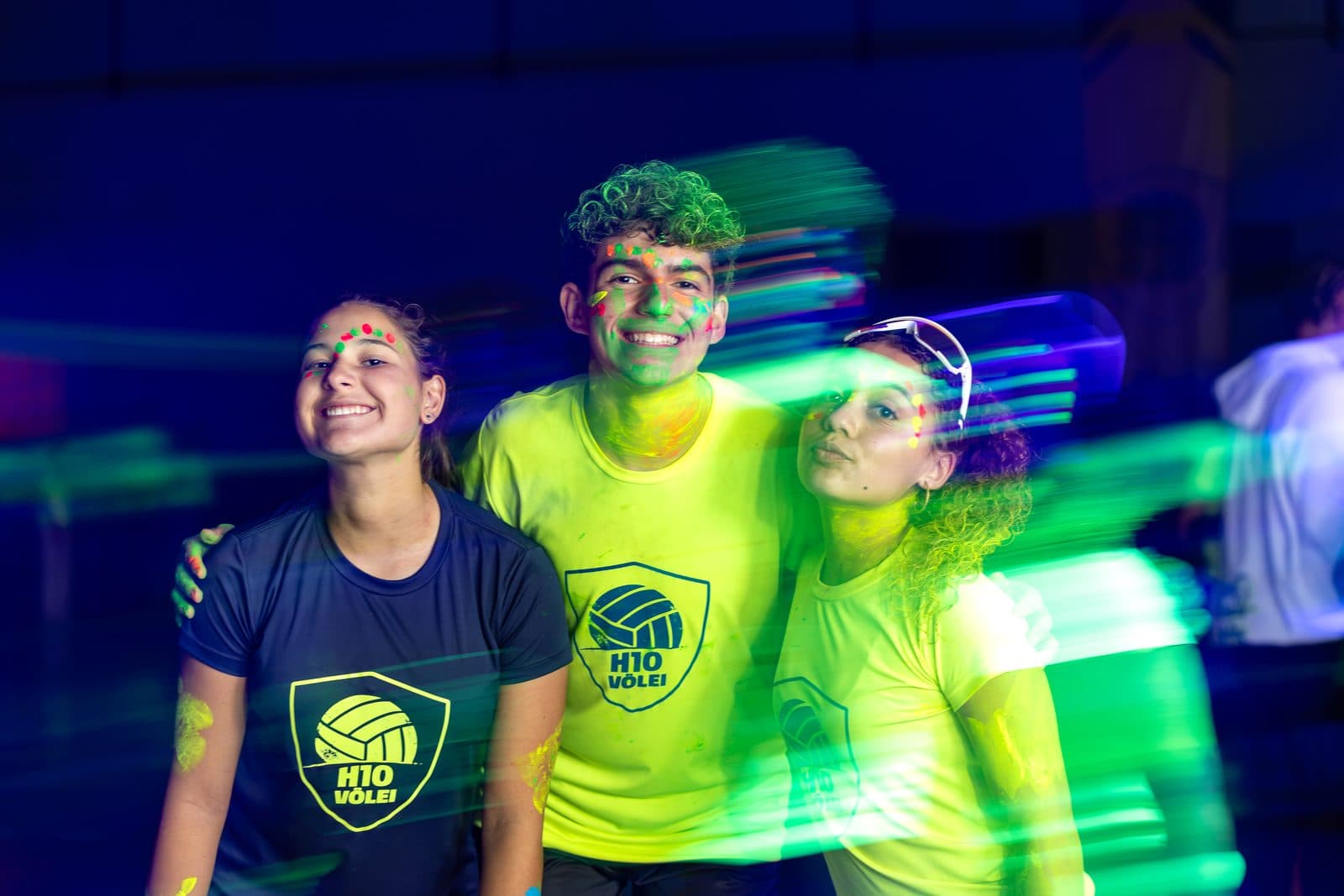 Trio de acampantes com pintura neon no rosto sob luz negra, rastros de luz verde atravessando o quadro