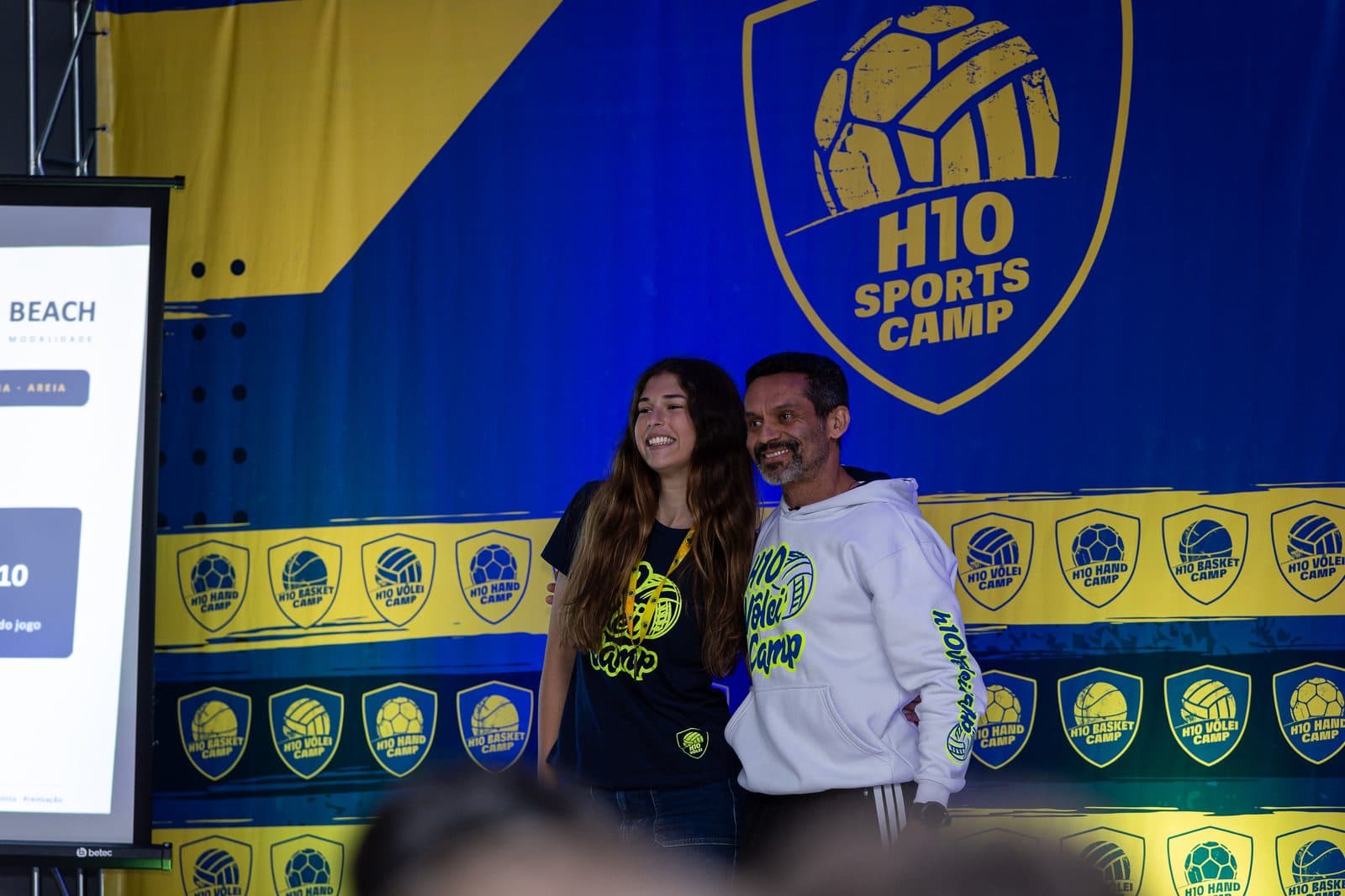 Acampante e integrante da equipe lado a lado no palco, diante do backdrop azul e amarelo H10 SPORTS CAMP