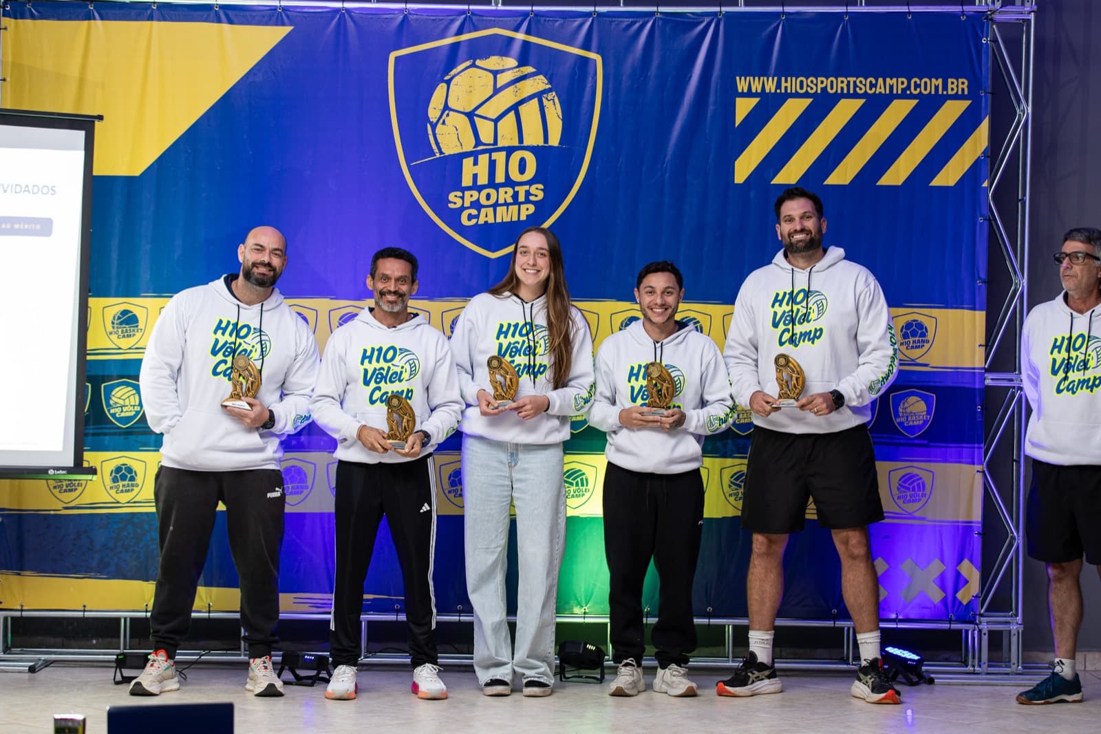 Equipe tecnica no palco com trofeus diante do backdrop H10 SPORTS CAMP sob luz roxa